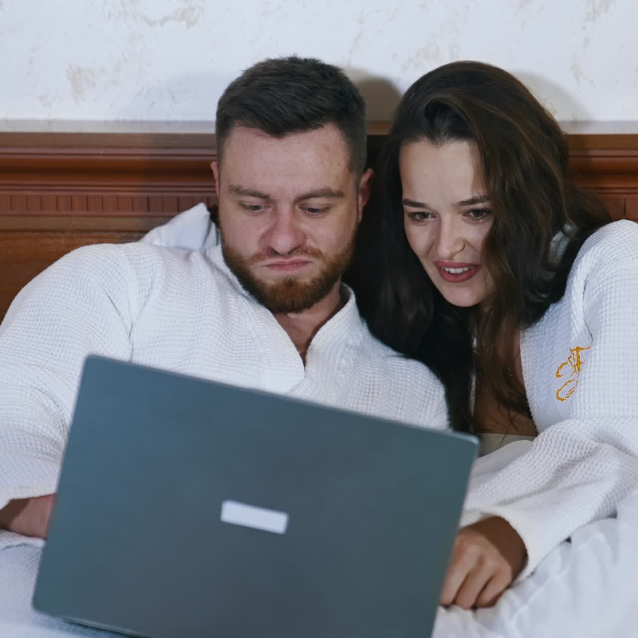 Young couple spend time together. Beautiful woman and her husband in white dressing gowns laying on bed and looking into the laptop