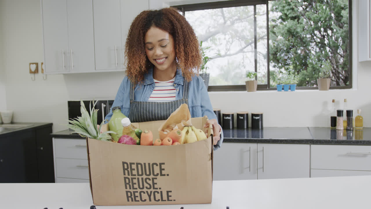 Happy biracial woman unpacking groceries from box with recycle text in kitchen, slow motion