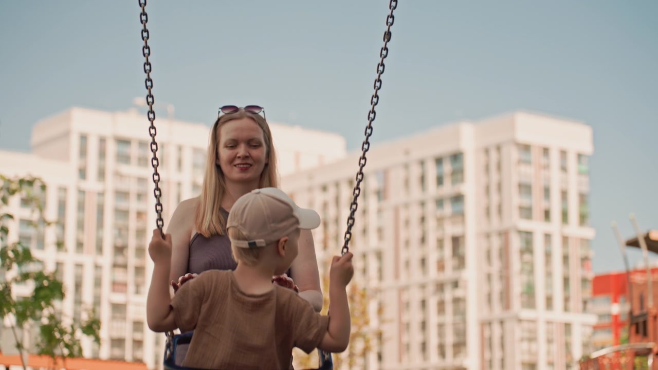 Una donna spinge delicatamente un bambino sull'altalena in un parco giochi all'aperto con edifici alti sullo sfondo. La scena cattura la loro interazione, mostrandoli mentre si godono il tempo insieme alla luce del giorno con una luce soffusa.