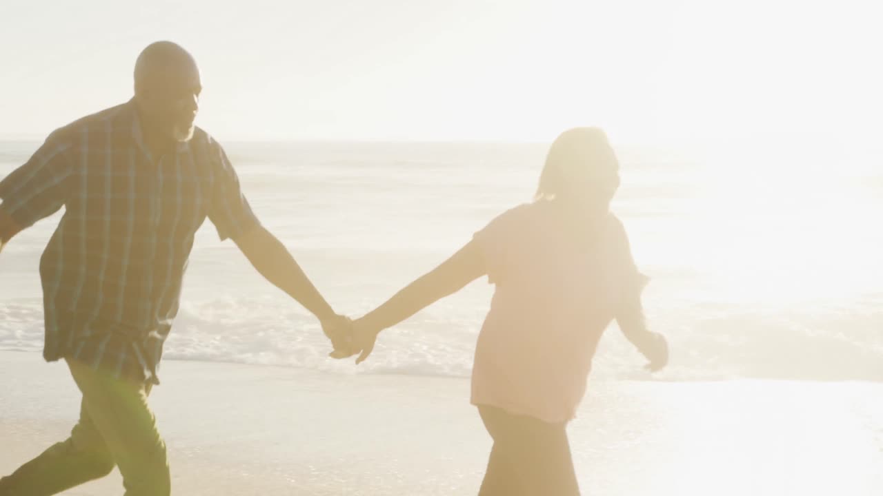 미소 짓는 아프리카계 미국인 부부가 손을 잡고  ⁇ 은 해변에서 달리고 있습니다.