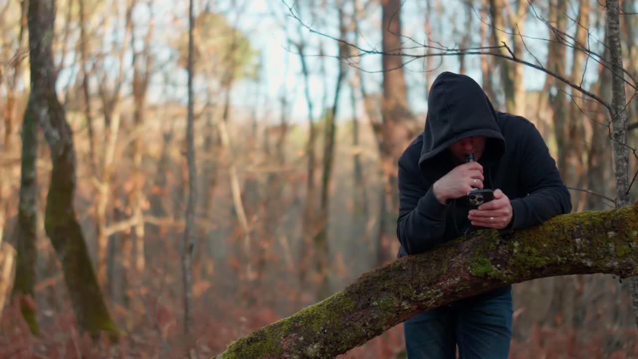Rallentatore. Giovane uomo con felpa nera con cappuccio che svapa sigaretta elettronica e scrive sullo smartphone nella foresta. Uomo che usa il cellulare e fuma sigaretta elettronica all'aperto tra gli alberi.