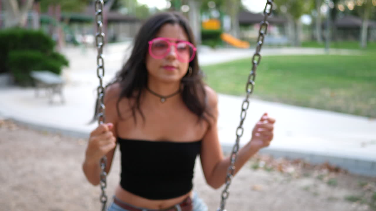 Primo piano di una graziosa ragazza ispanica che gioca sull'altalena del parco giochi indossando occhiali da sole rosa retrò al rallentatore
