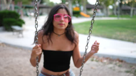 cerrar a una linda chica hispana jugando en el columpio del patio de recreo con gafas de sol rosas retro en cámara lenta