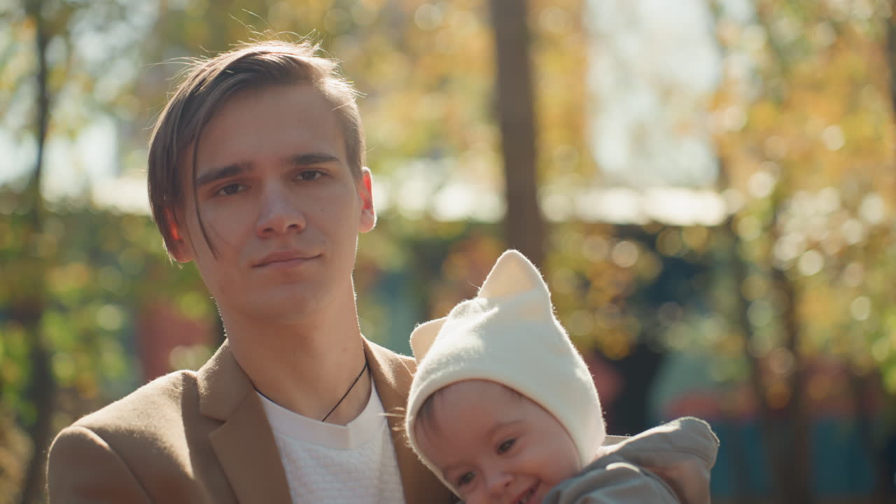 Ung far holder baby tæt i parken, rolig beskyttende stemning med blødt efterårslys. Stoisk far i blazer med spædbarn, der putter sig mod skulderen, filmiske varme toner og lav dybdeskarphed, øm familie