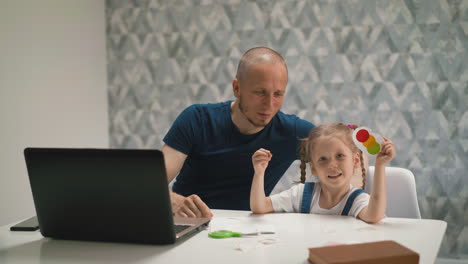 cute-girl-shows-colorful-craft-near-smiling-father-at-table