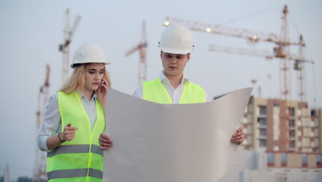 Zwei-Bauarbeiter-Mit-Zeichnungen-Stehen-In-Helmen-Und-Westen-Vor-Dem-Hintergrund-Im-Bau-Befindlicher-Gebäude,-Eine-Frau-Telefoniert-Mit-Einem-Kunden.