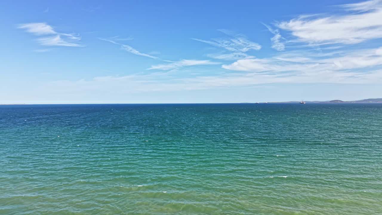 Ampia vista dell'oceano con cielo sereno a mezzogiorno, che mostra acqua calma e barche lontane al largo della costa.