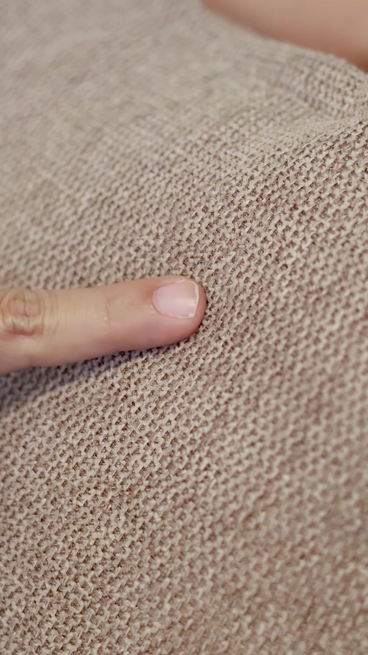 A hand points at textured beige upholstery fabric in a bright, indoor setting.