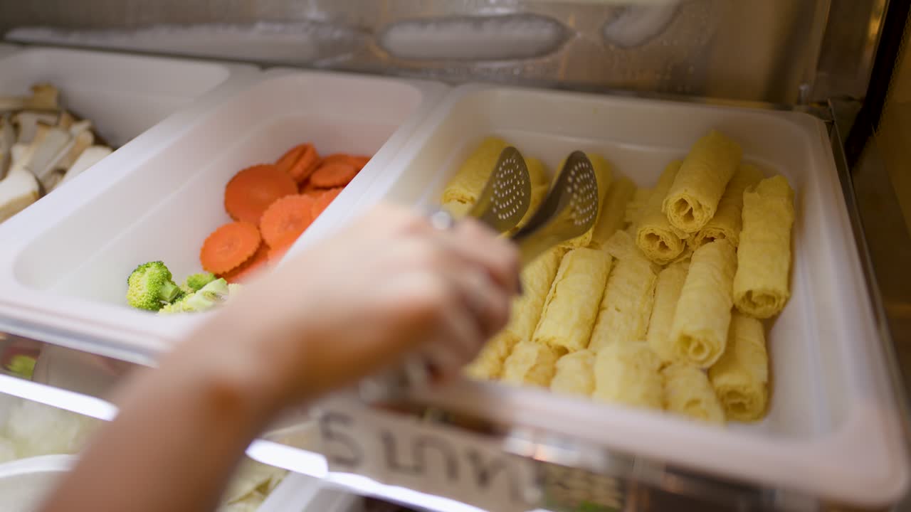 Hand uses tongs to pick tofu skin rolls from buffet tray under soft, warm lighting