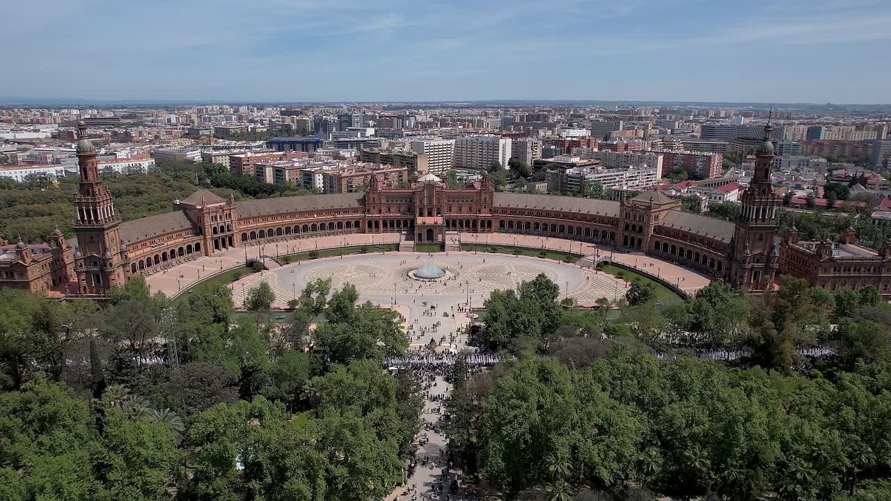 Wide zoom in aerial over Plaza de Espana in the Parque de María Luisa, Seville showing semicircular neoclassical Mudejar building, central fountain and visitors crossing sunlit expansive park square