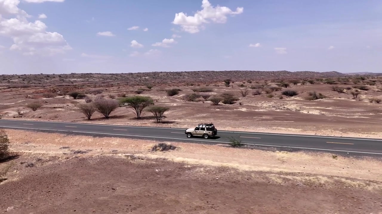 Ripresa aerea con drone di un SUV che percorre una strada asfaltata tortuosa attraverso un vasto paesaggio desertico, arido e roccioso con vegetazione rada sotto un cielo pallido