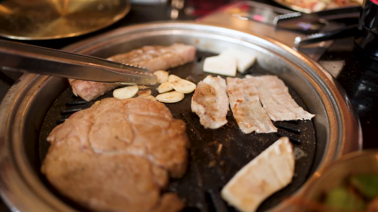 Tongs grasp sliced garlic on a hot grill with pork and vegetables, warm indoor lighting