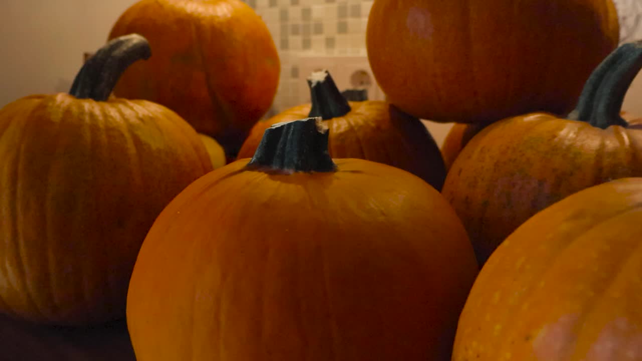 Close up footage of pumpkins piled on top of each other while being placed on a kitchen table indoors under warm lights. Pumpkins have gorgeous orange color and stems with light reflecting off them.