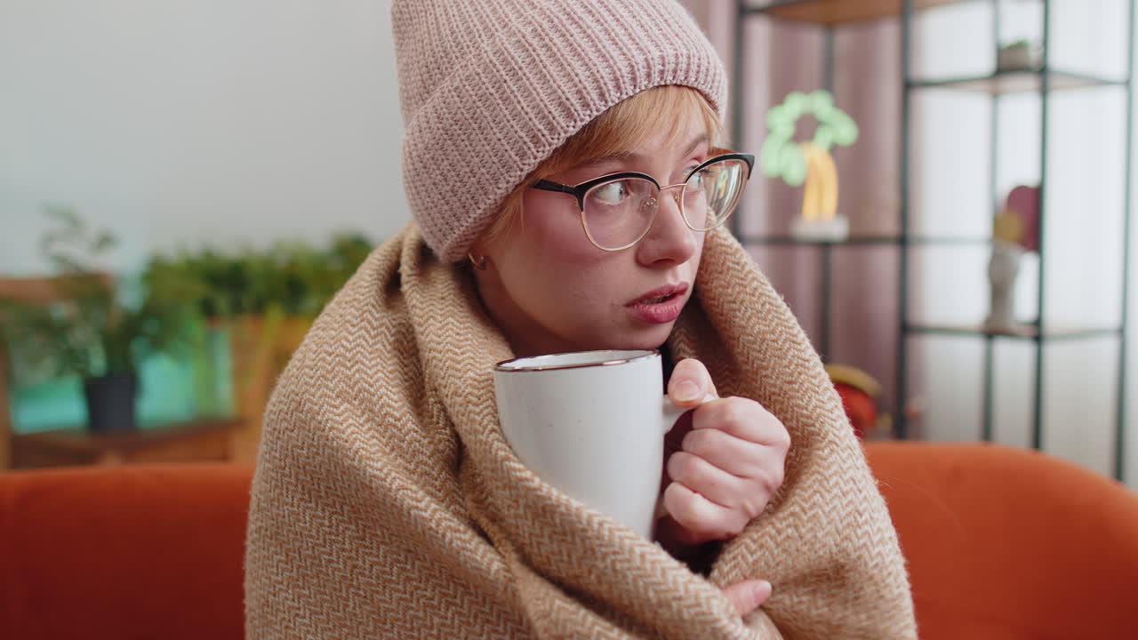 Sick woman girl wear hat wrapped in plaid sit alone shivering from cold on sofa drinking hot tea