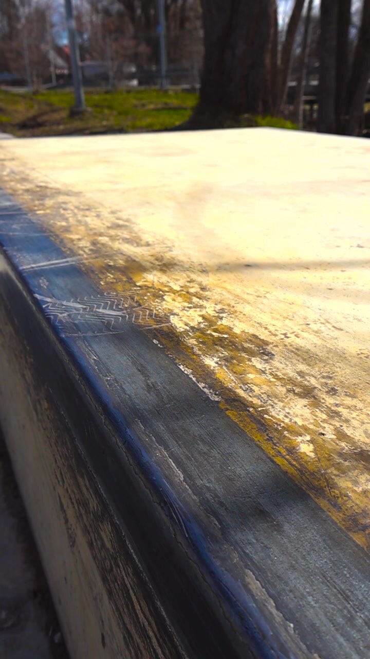 Primo piano che scorre lungo un bordo di cemento usurato e graffiato con un coping in metallo nero in uno skatepark esterno, mostrando segni di utilizzo e usura dovuti a grind e slide con lo skateboard