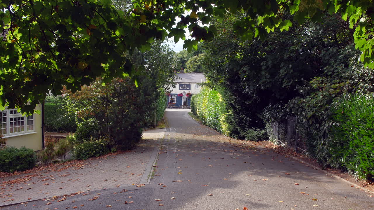 A quiet path lined with trees and hedges leads to a quaint pub named "The Railway." Dappled sunlight filters through the leaves, with fallen autumn leaves scattered along the paved walkway