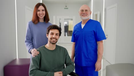 retrato de un feliz hombre moreno con paja en una chaqueta verde que se sienta en una silla de ruedas después de ser herido cerca de una chica morena y un experimentado médico hombre confiado en un uniforme azul en una clínica moderna