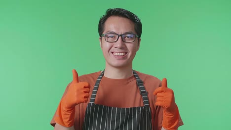 primer plano de una ama de llaves asiática con un delantal sonriendo y mostrando los pulgares hacia arriba mientras está de pie en el estudio de pantalla verde