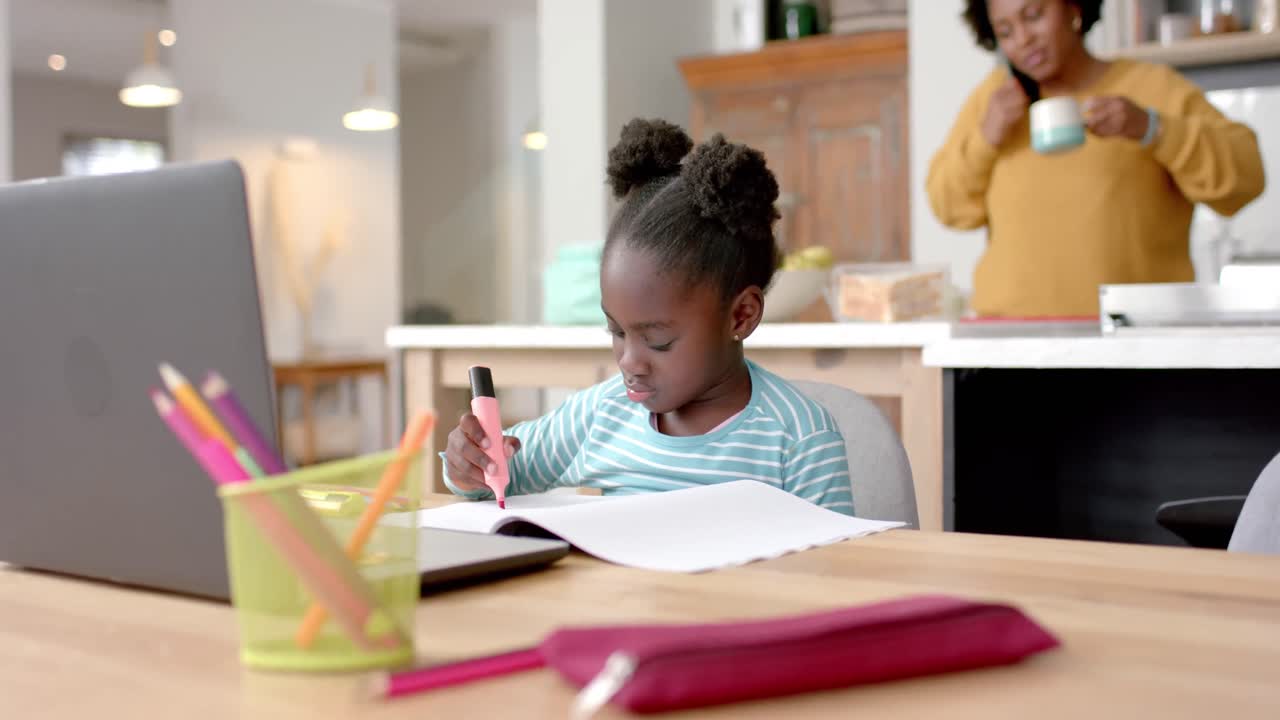 African american girl learning online at table using laptop with mother in kitchen, slow motion