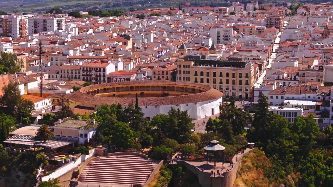Filmato drone che si alza e si sposta lateralmente sopra la famosa arena di Ronda, tetti in terracotta fitti, strade alberate e una grande facciata di hotel con la catena della Sierra Nevada sullo sfondo
