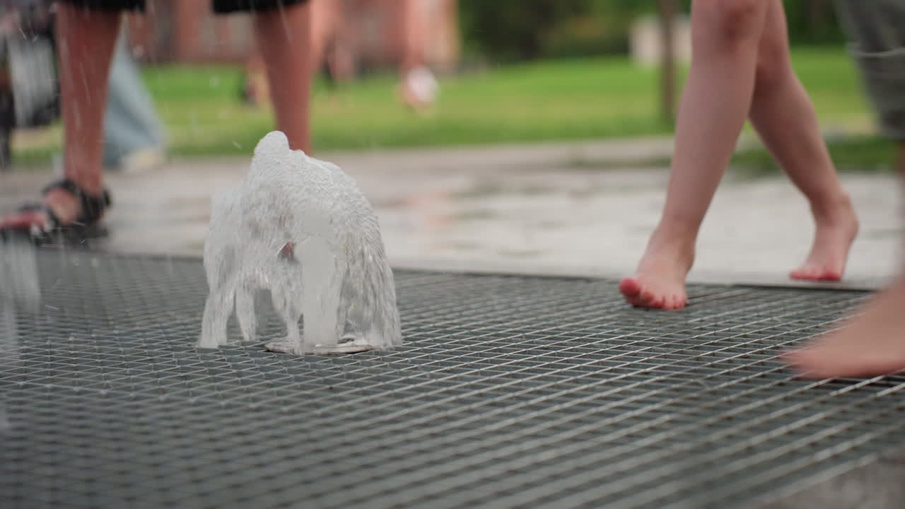 Playful child using leg with trouser folded to close fountain jet while girl reaches with hand touching water, blurred people in background, summer park setting showing curiosity and interaction