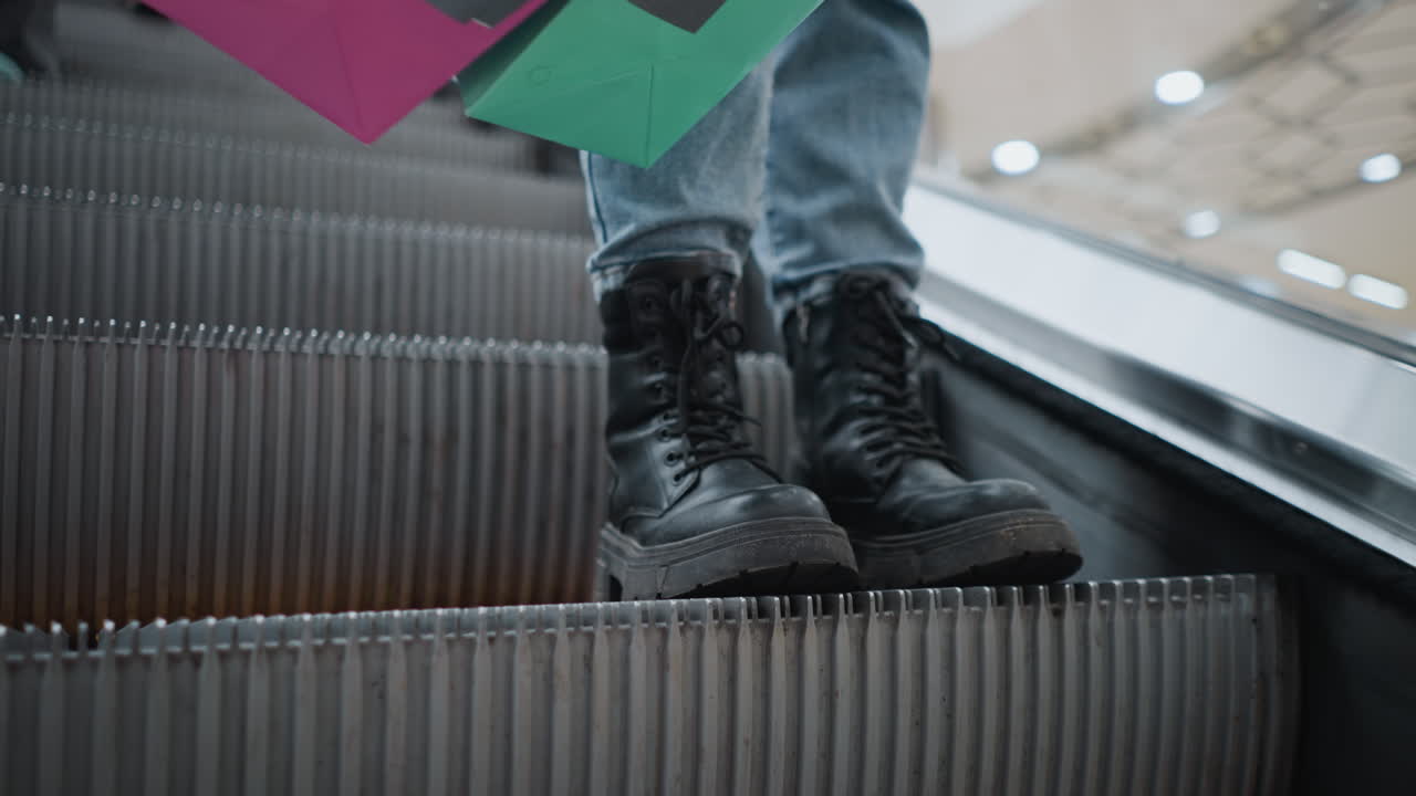 close-up de pernas vestindo botas pretas e jeans de pé perto da corrimão da escada rolante enquanto carrega um saco de compras colorido com visão parcial de outra pessoa no fundo