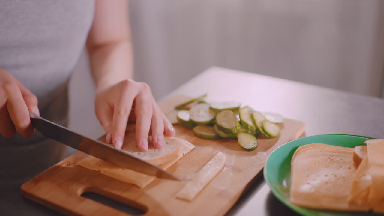 Close up of person cutting bread slice with cheese on wooden board beside fresh cucumber slices and extra cheese on plate, preparing simple sandwich ingredients in bright kitchen atmosphere