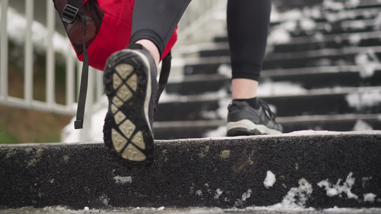 primer plano de las piernas en zapatillas de deporte caminando por una escalera nevada mientras llevan una mochila roja, escaleras cubertas de nieve con barandilla de hierro al lado y calzado deportivo