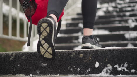 close-up of legs in sneakers walking up snowy staircase while carrying red backpack, snow-dusted stairs with iron railing by the side and athletic footwear