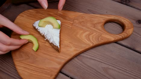 a woman's hand cuts a piece of avocado in half with a steel knife and places it on a buttered sandwich. then he puts dill on top. preparing breakfast