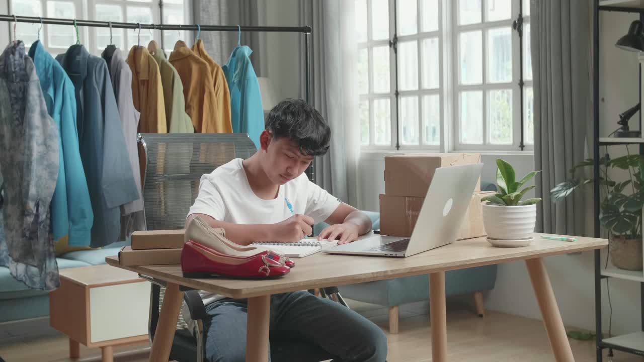 Asian Man Online Seller Looking At Computer Screen And Writing In The Notebook While Selling Clothes At Home