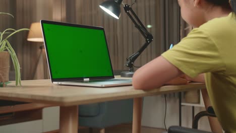 back view of asian kid girl sitting in a wheelchair while using laptop computer with mock up green screen and note in book at home
