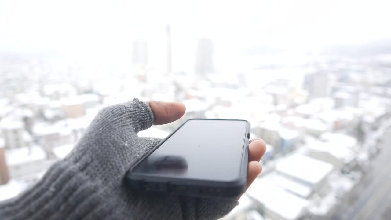 Hånd i en grå hanske som holder en smarttelefon mot en snødekt bybakgrunn