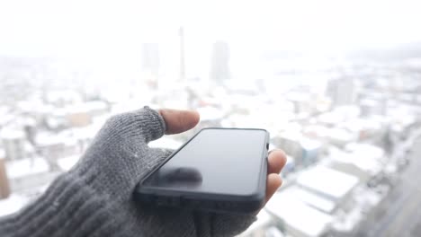 hand in a grey glove holding a smartphone against a snowy city background