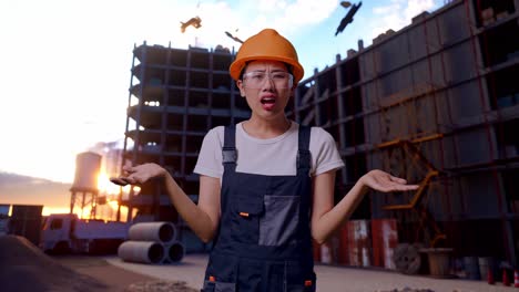 asombrada trabajadora asiática con gafas de protección y casco de seguridad diciendo por qué y de pie con dudas en el sitio de construcción