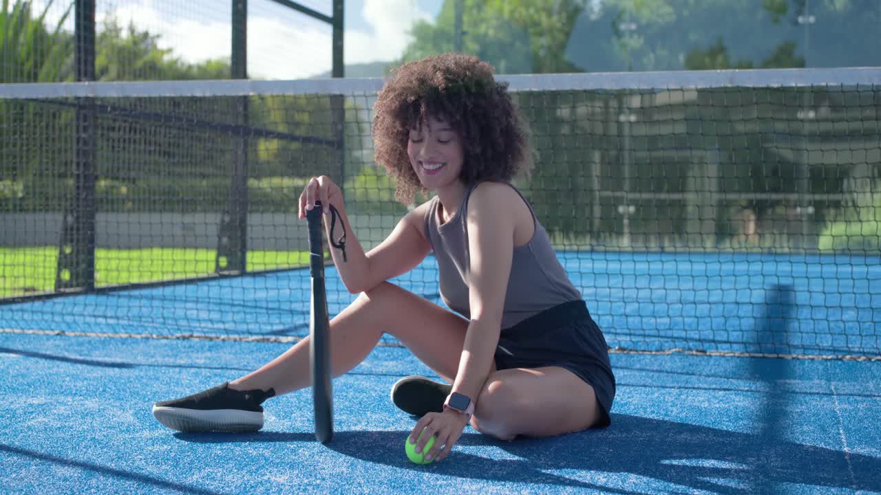 Voksen kvinne sitter på blå bane etter en pause, tar opp og ruller ballen, og forbereder seg på å spille med racket.