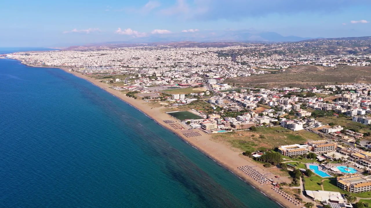 Luftfoto fra drone af en lang sandstrand med strandresorthoteller og pools, med byen langs kysten i baggrunden, Ammoudara, Grækenland.