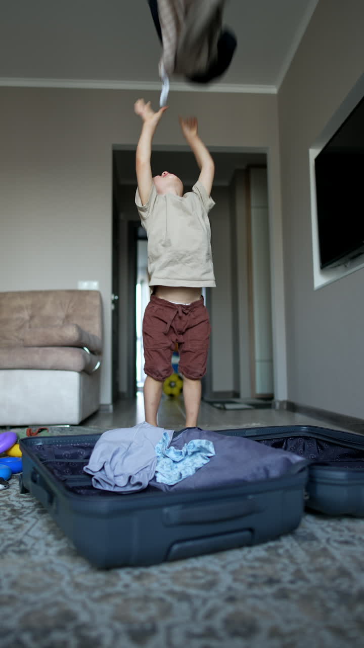 Cheerful baby boy takes some clothes out of the suitcase and throws them up. Happy kid rejoices and claps hands actively. Vertical video.