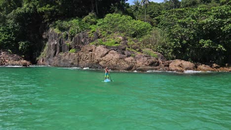 low angle aerial drone bird's eye view of man exercising on a sup paddle board in turquoise tropical clear waters, rocky coastline and tropical jungle forest in thailand