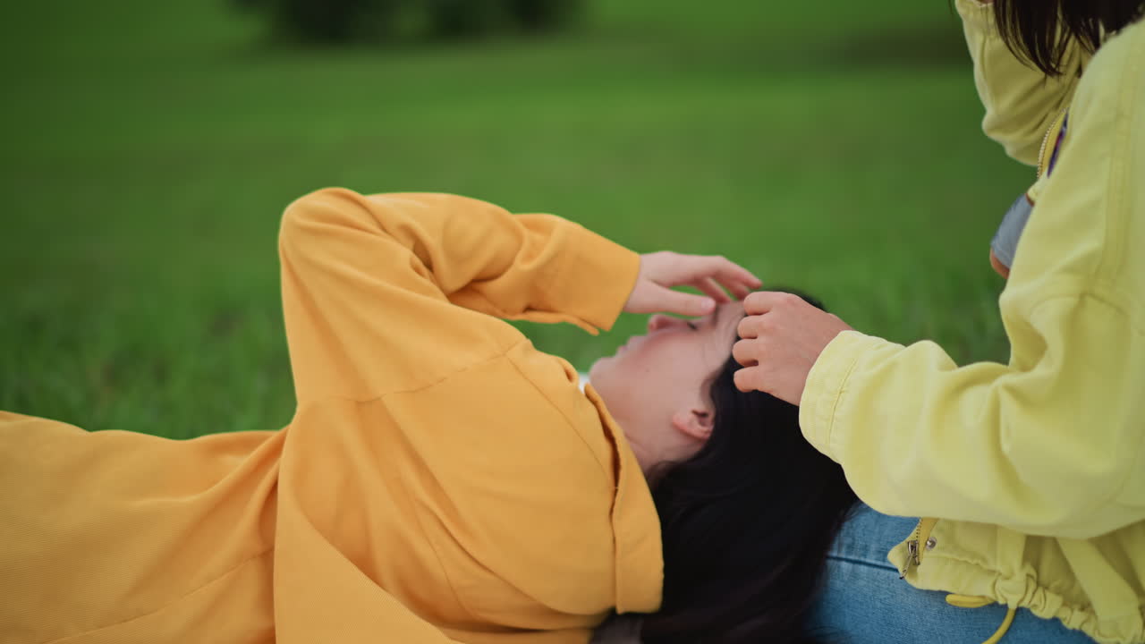 asian woman lying on grass, friend reaching over to gently cover eyes and stroke hair, warm yellow sweater and denim, soft picnic blanket, green park background, afternoon light casting calm mood