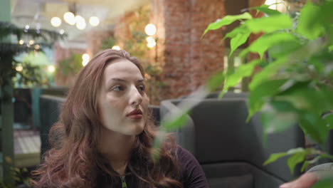 woman with curly hair in black outfit walks inside cozy green-themed lounge and gently reaches toward leafy plant while surrounded by brick walls, warm lighting, indoor trees, and green furniture