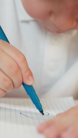 Concentrated little boy draws on notebook page with blue marker at table closeup. Toddler student works at art lesson in preschool classroom