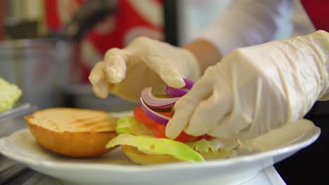 jefe con guantes poniendo vegetales en un bollo para hamburguesa