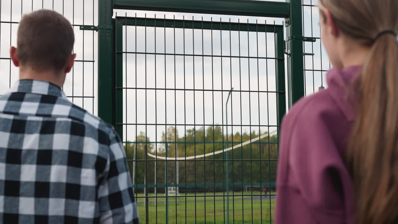 primer plano de jóvenes entrando en la cancha de voleibol mientras el entrenador en camisa a cuadros abre la puerta verde, con el campo de fútbol y los árboles en el fondo, simbolizando la preparación para el entrenamiento deportivo