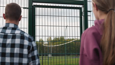 close-up of young people entering volleyball court as coach in plaid shirt opens green gate, with football field and trees in background, symbolizing preparation for sports training