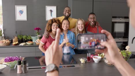 Biracial-man-using-smartphone-and-taking-picture-of-diverse-friends-in-kitchen,-slow-motion