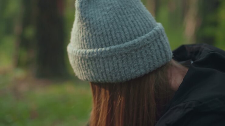 Close up of woman wearing beanie smiling warmly while conversing outdoors in forest, sunlight softly casts over her face highlighting cheerful expression and relaxed mood against blurred background