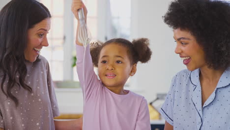 familia con dos madres vestidas con pijama haciendo panqueques matutinos en la cocina en casa con la hija