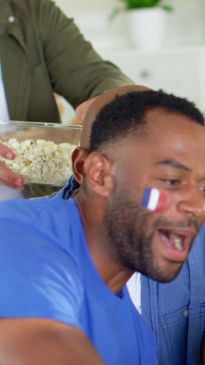 Vertical video: Cheering man in blue tee flag paint on TV at home, pal taking popcorn, copy space