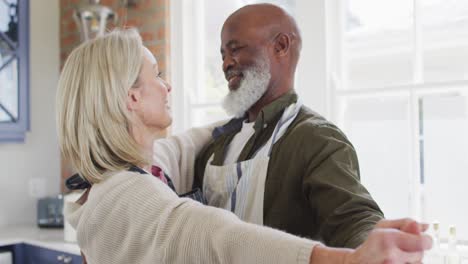 Mixed-race-senior-couple-wearing-aprons-dancing-together-in-the-kitchen-at-home